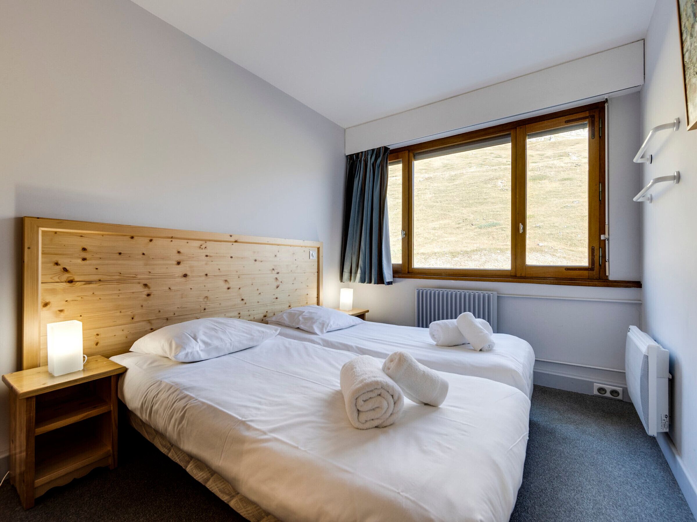 A hotel room with a wooden headboard, two beds, a nightstand with a lamp, a window with curtains, and a radiator.