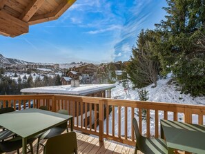 A wooden deck with a table and chairs, overlooking a snowy landscape with buildings and trees.