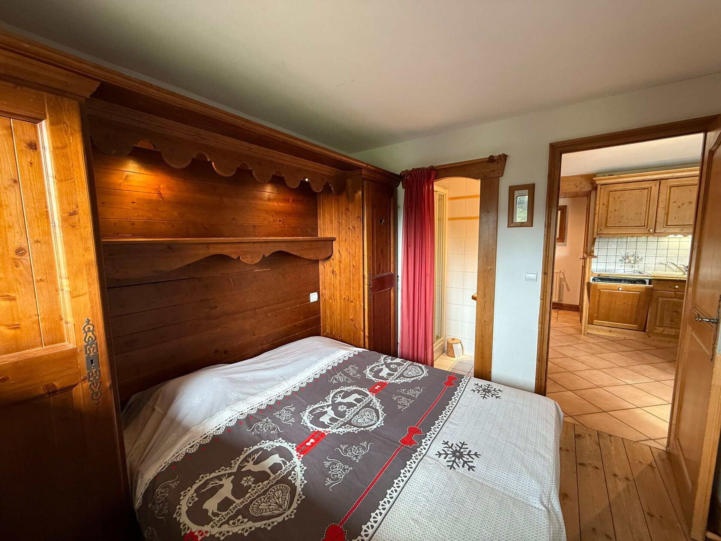 A wooden bedroom with a bed, a wooden headboard, and a view of a kitchen through an open door.