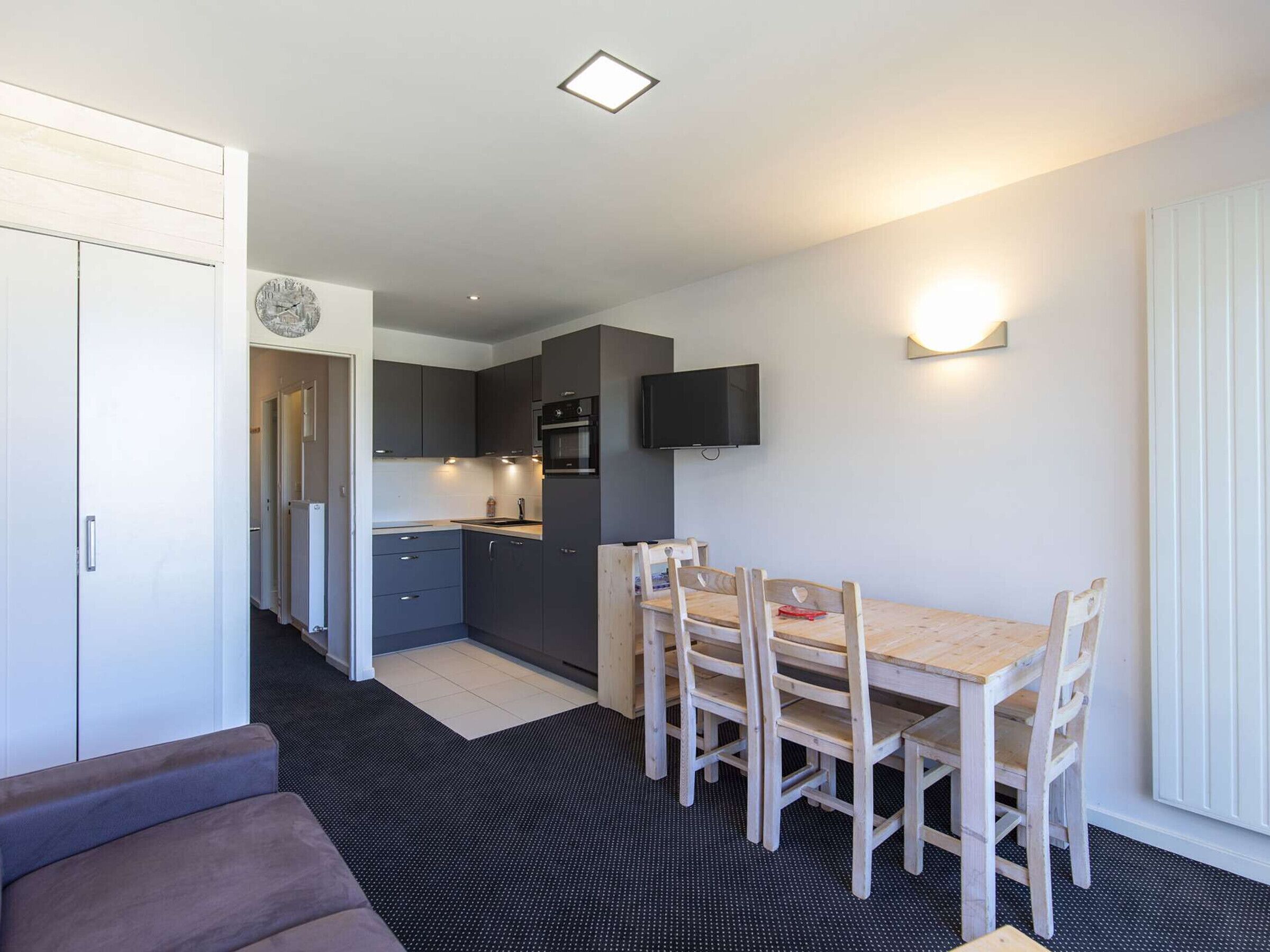 A compact kitchen with modern appliances, a dining area with a wooden table and chairs, and a flat-screen TV mounted on the wall.