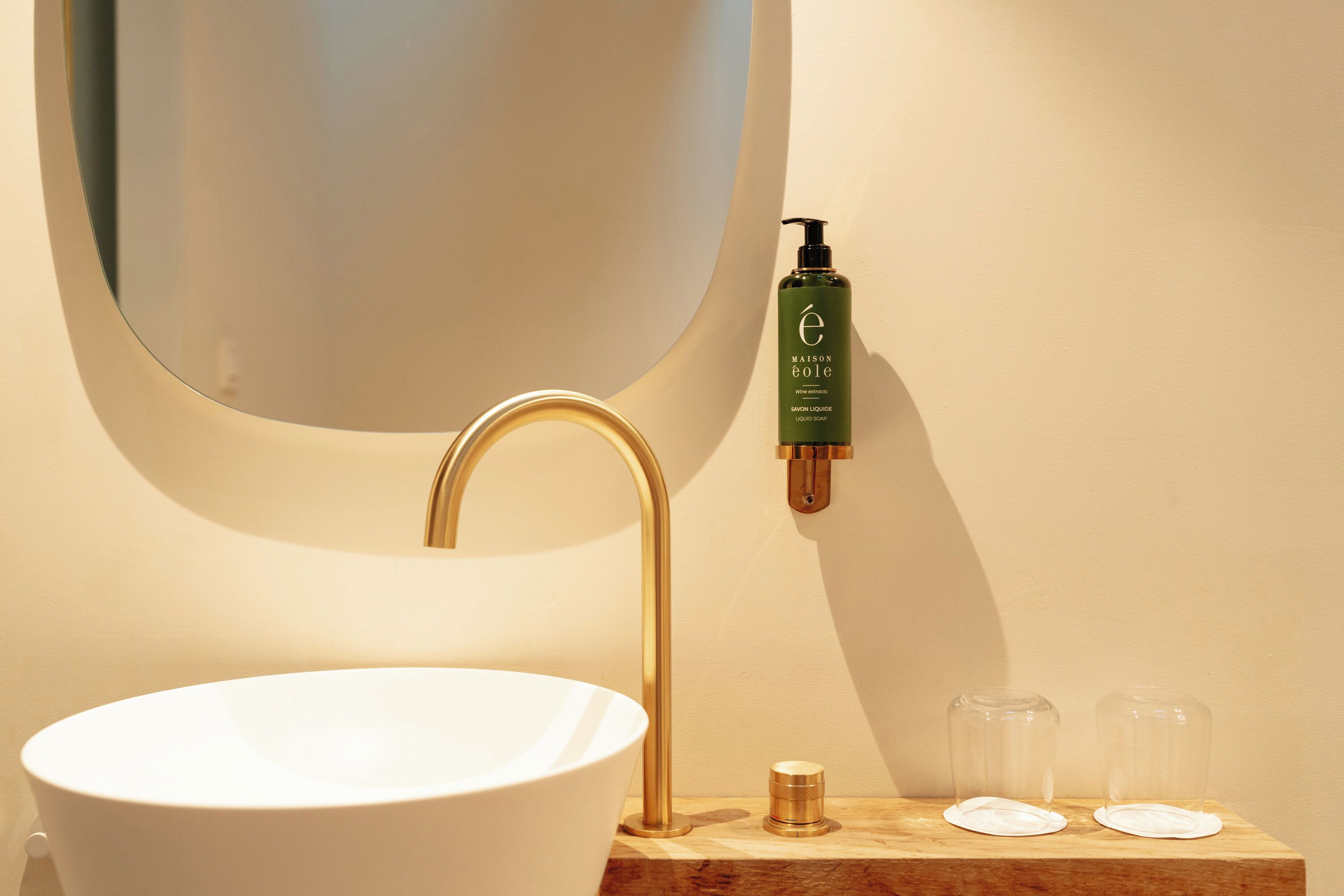 A bathroom with a white sink, a gold faucet, a green bottle of Maison eole soap, and two clear glass containers on a wooden shelf.