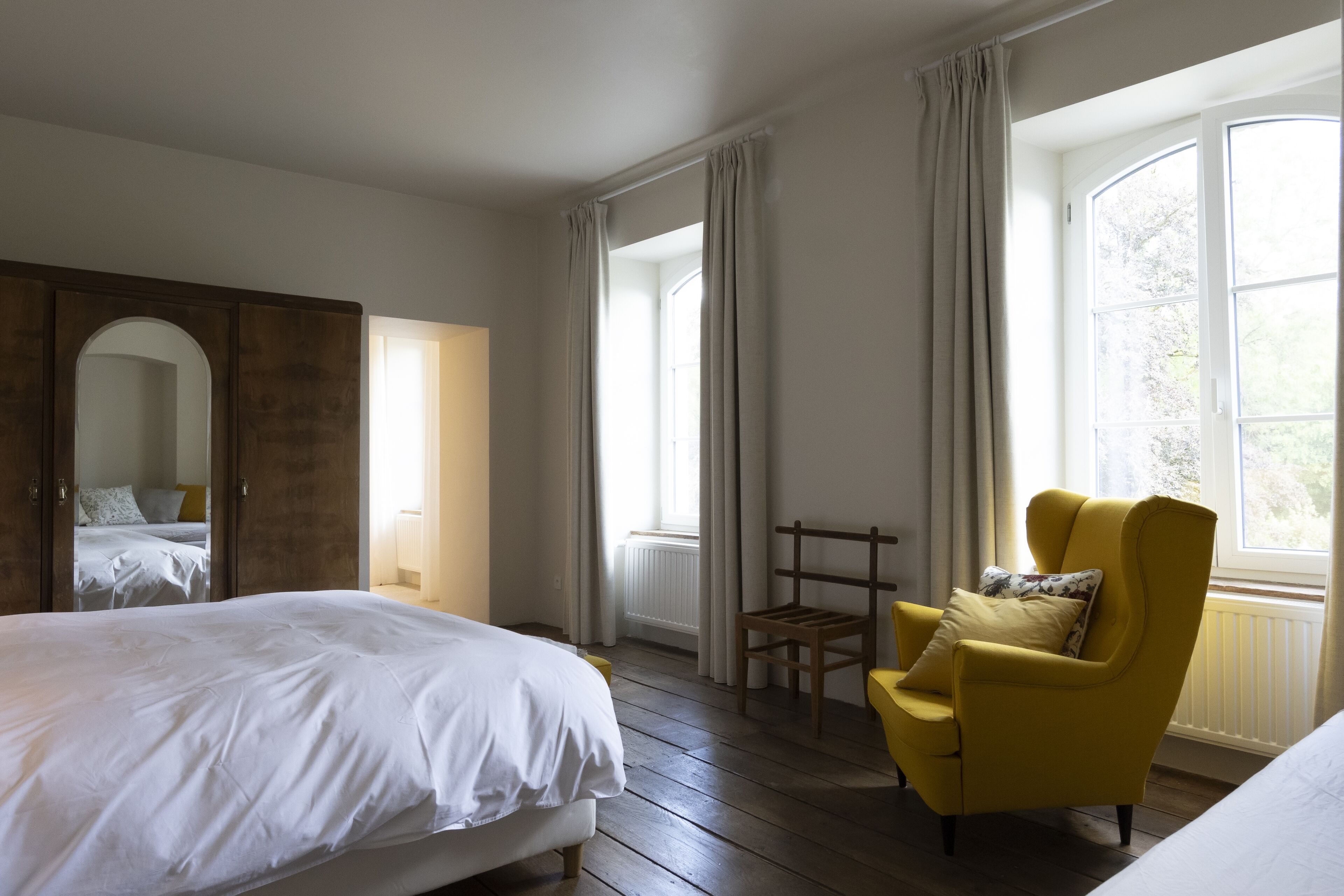A bedroom with a large bed, a yellow armchair, and a window with curtains.