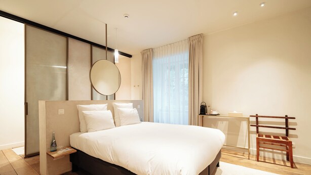A modern bedroom with a large bed, bedside table, mirror, and a wooden chair. - Château du Pont d'Oye (Habay)