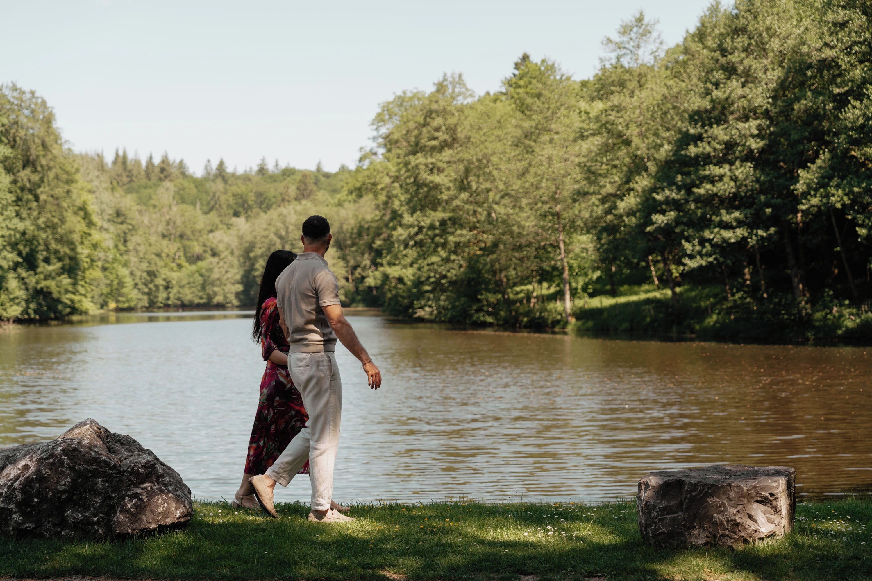 A couple walking by a lake with trees in the background.