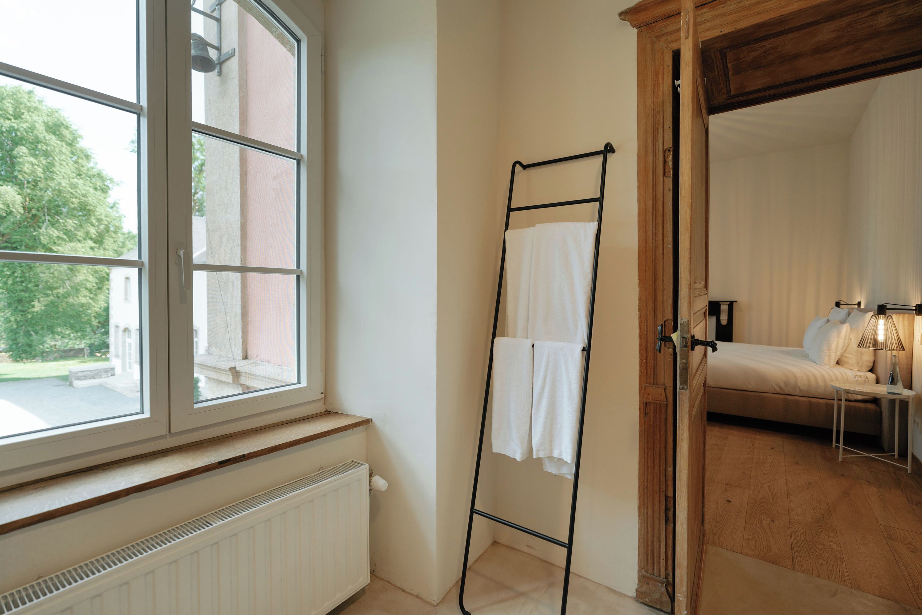 Una habitación con ventana, radiador, suelo de madera, cama, mesita de noche, lámpara y un perchero para toallas.