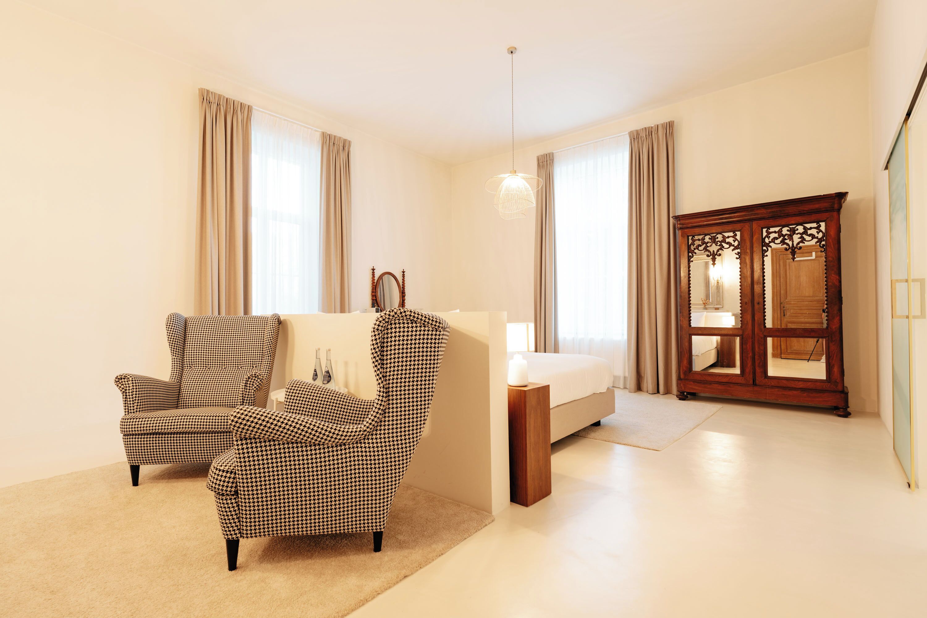 A bedroom with a bed, a wooden wardrobe, a checkered armchair, a small table, and a hanging light fixture.