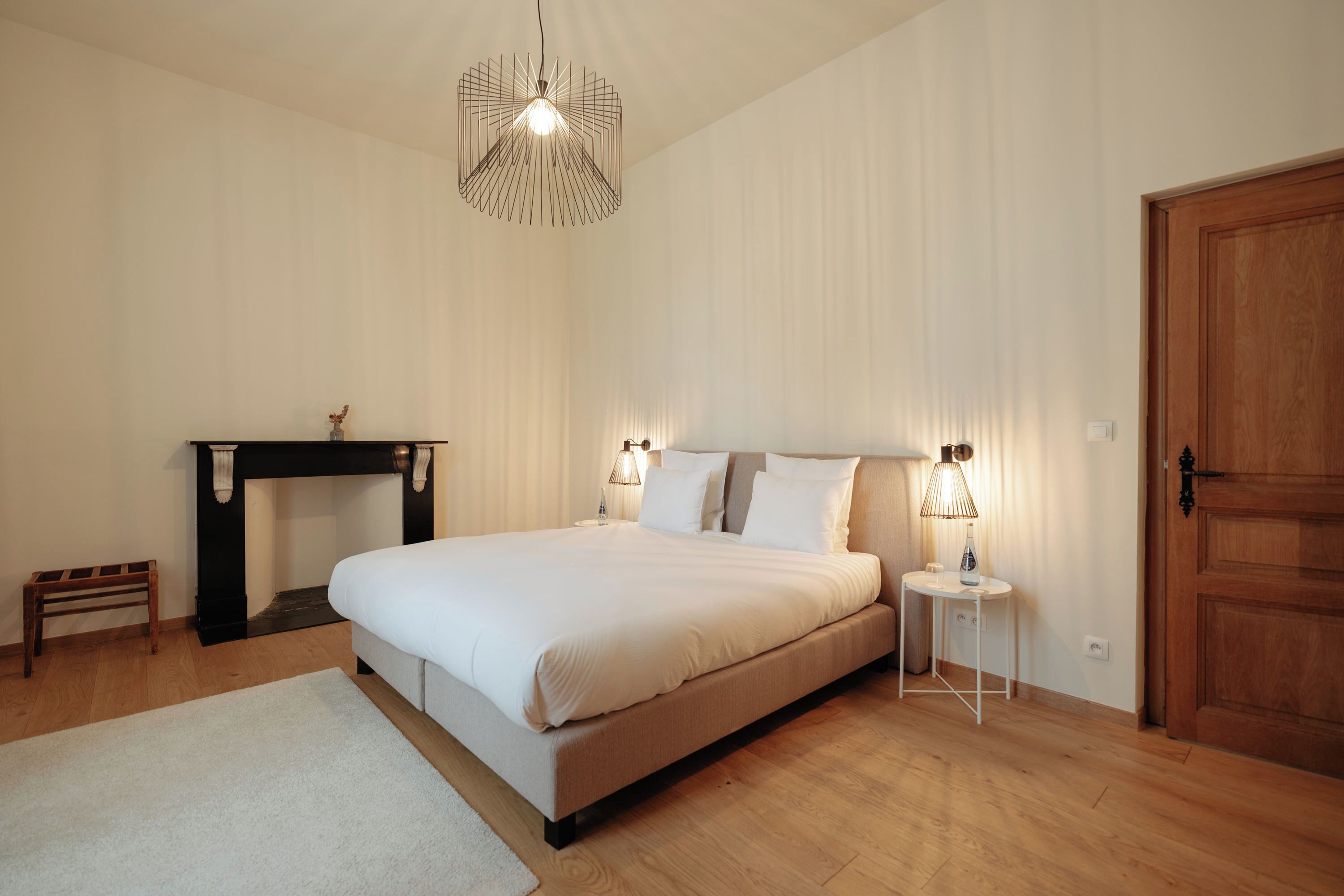 A bedroom with a bed, bedside lamps, a nightstand, a wooden bench, a fireplace, and a door.