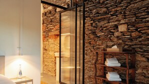 A modern bathroom with a stone wall, a glass shower enclosure, a wooden shelf with towels, and a small bedside table with a bottle. - Château du Pont d'Oye (Habay)