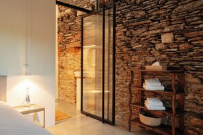 Ein modernes Badezimmer mit Steinwand, einer Duschkabine aus Glas, einem Holzregal mit Handtüchern und einem kleinen Nachttisch mit einer Flasche.