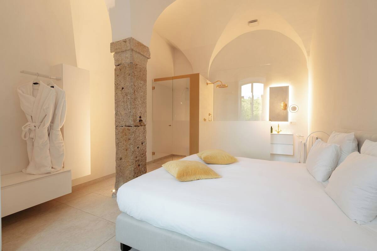 Ein Schlafzimmer mit einem großen Bett, einer Steinsäule, einem Kleiderständer und einem durch einen Rundbogen sichtbaren Badezimmer.