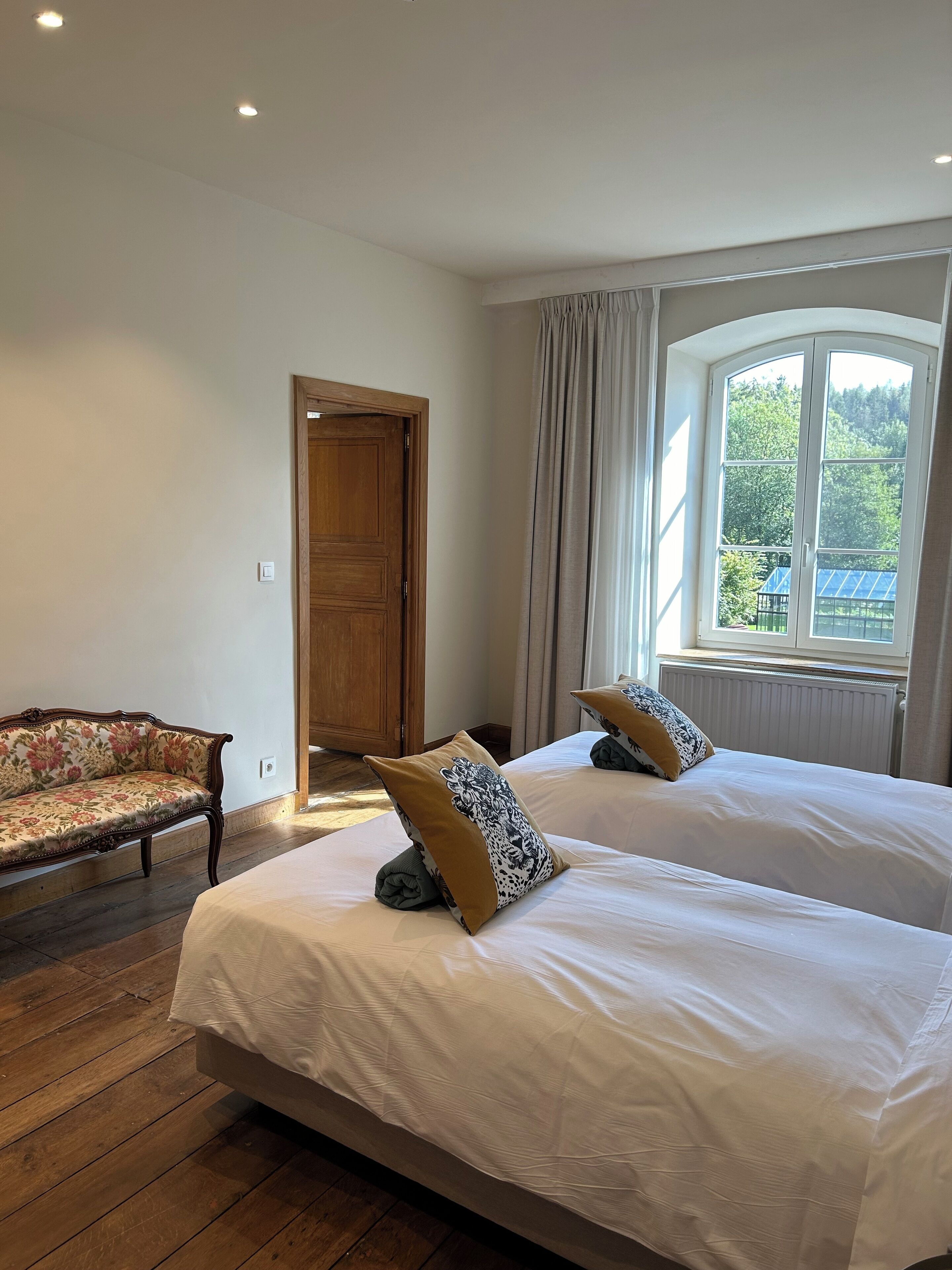 A bedroom with two beds, a wooden door, a sofa, and a window with a view of greenery.
