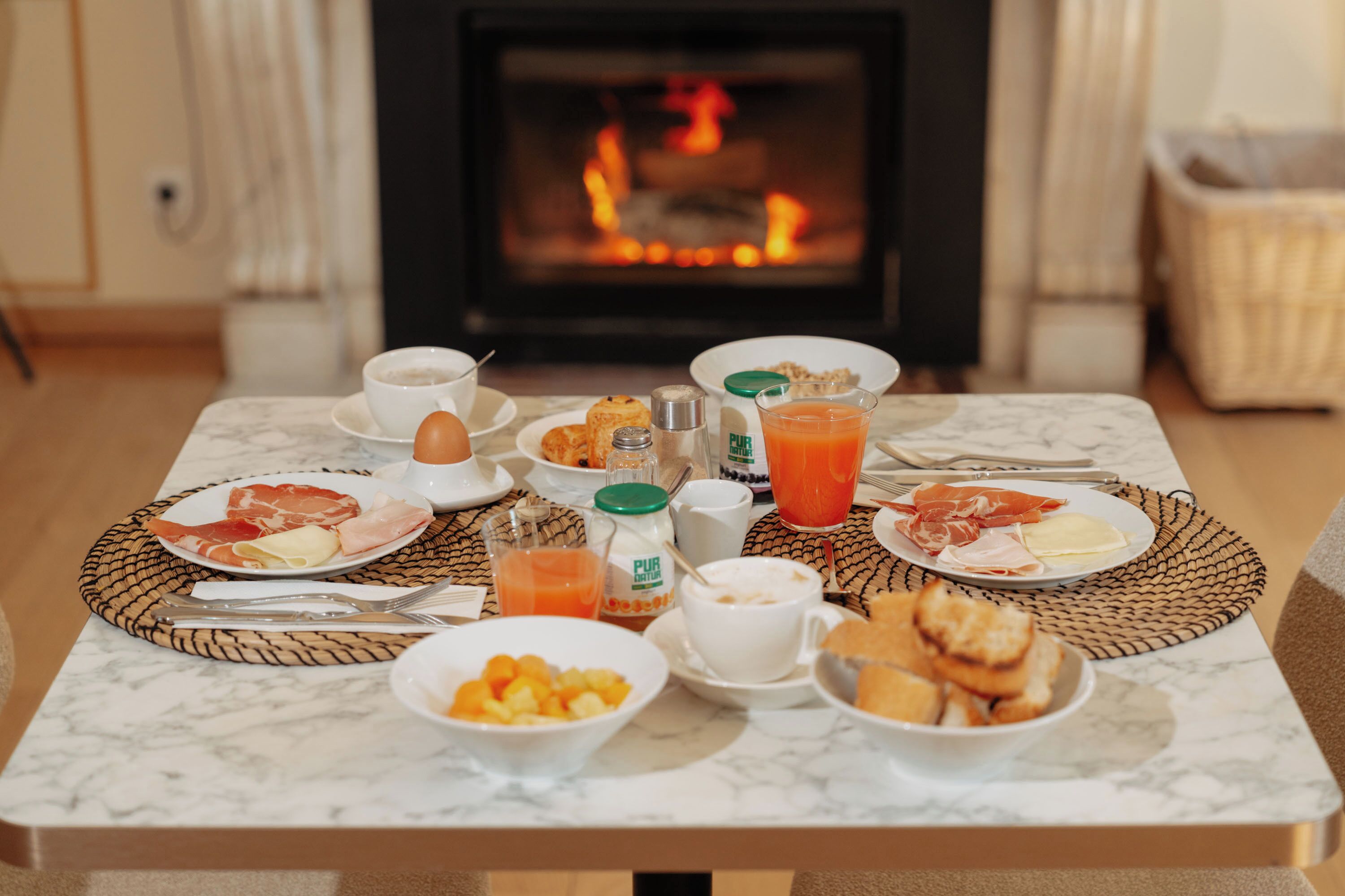 A breakfast table with plates of food, a jug of orange juice, and a view of a fireplace.