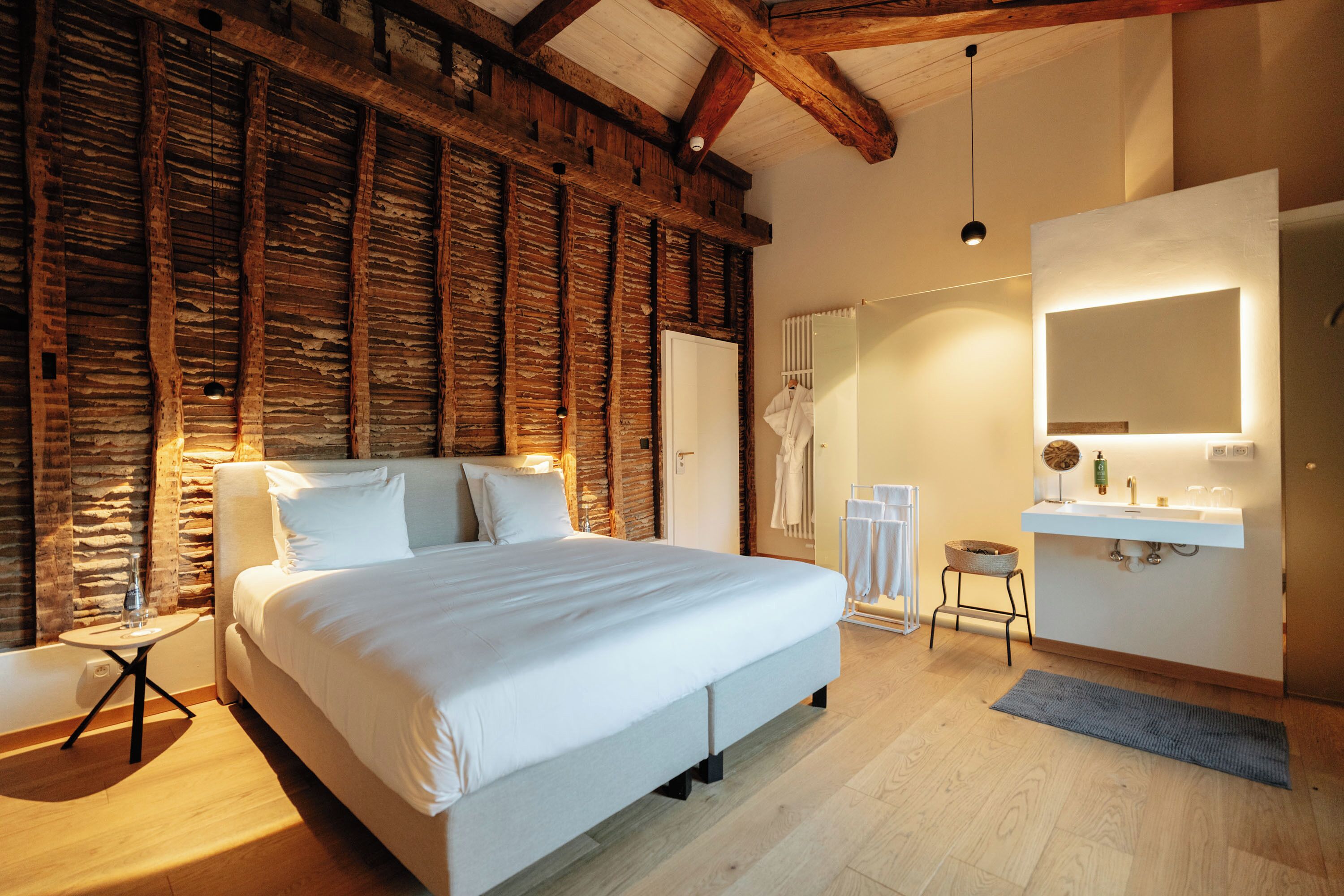 A bedroom with a large bed, a wooden ceiling, a white wall with a mirror, and a small stool.