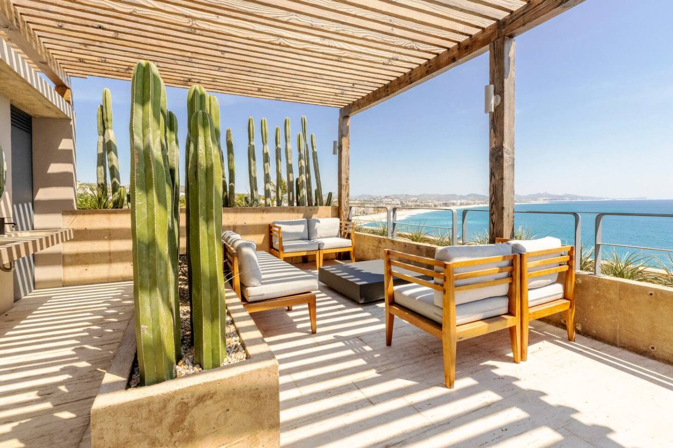 A sunlit outdoor seating area with wooden furniture and cacti, overlooking a coastal view.