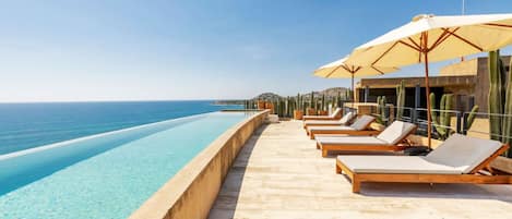 An infinity pool with lounge chairs and umbrellas, overlooking a clear blue sea.