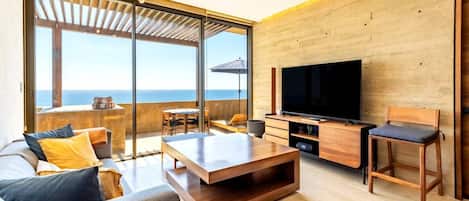 A modern living room with a large TV, wooden furniture, and a view of the sea.