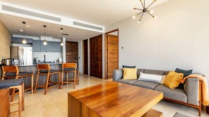 A modern living room with a wooden coffee table, a grey sofa with yellow cushions, and a kitchen area with wooden cabinets and bar stools. - Panoramic Ocean View - Hot Tub (San José del Cabo)