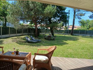 A backyard with a wooden deck, wicker furniture, a table with chairs, and a lawn.