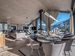 A modern living room with a dining area, large windows, and a view of snow-capped mountains.