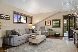 A modern living room with a grey sofa, a matching ottoman, a side table, and a chair. There are framed pictures on the walls, a window with a view, and a staircase leading to an upper level. - Private hot tub, large home, close to downtown (Red Lodge)