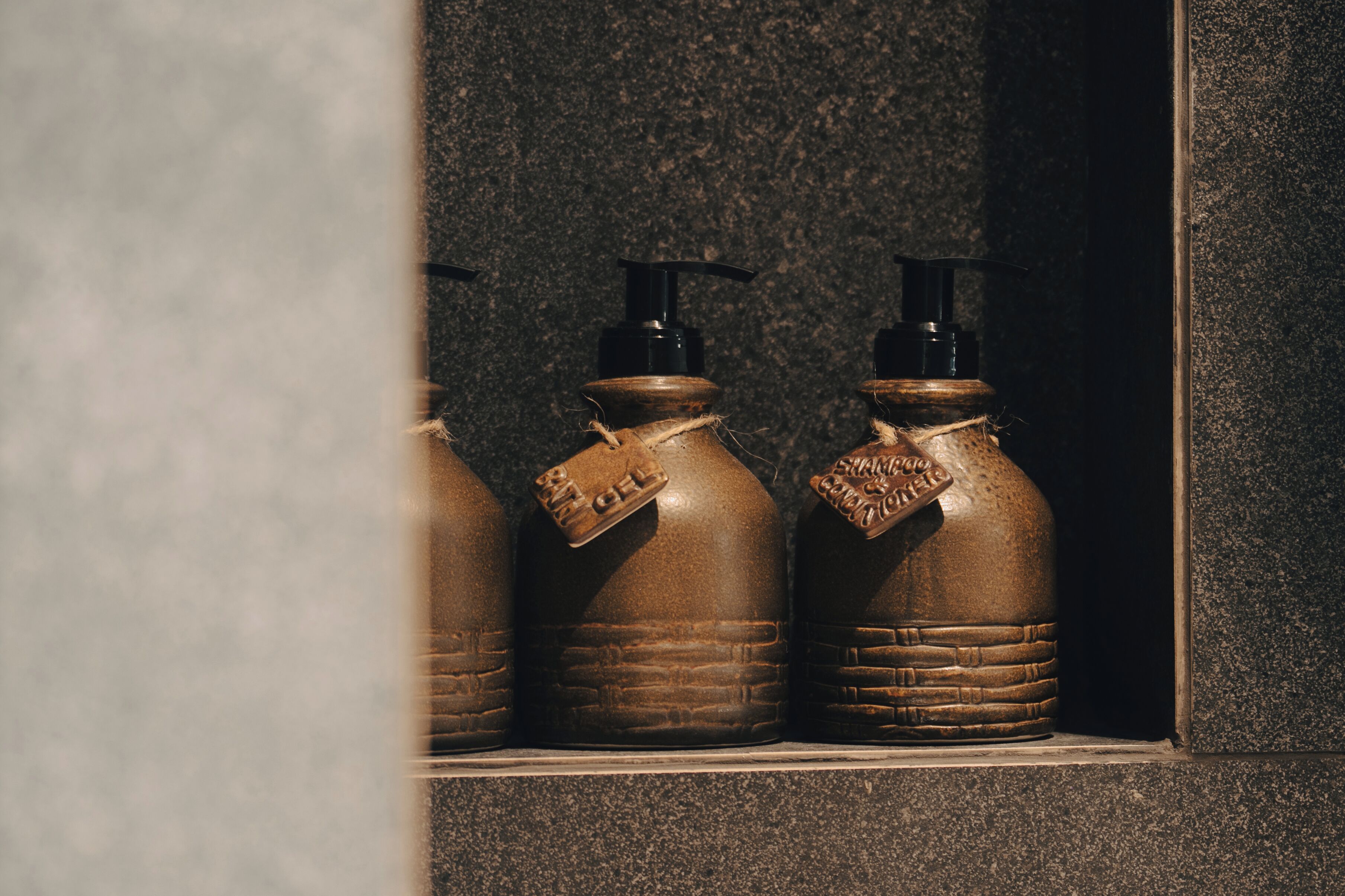 Tres botellas marrones con dosificadores negros y etiquetas en un nicho de pared de piedra.