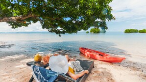 Une scène de plage avec une personne allongée sur une chaise longue sous un arbre, un kayak rouge et un fruit jaune sur le sable.