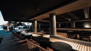 Un espace bar extérieur avec des tables et des chaises en bois, de grandes colonnes et une vue sur la mer.