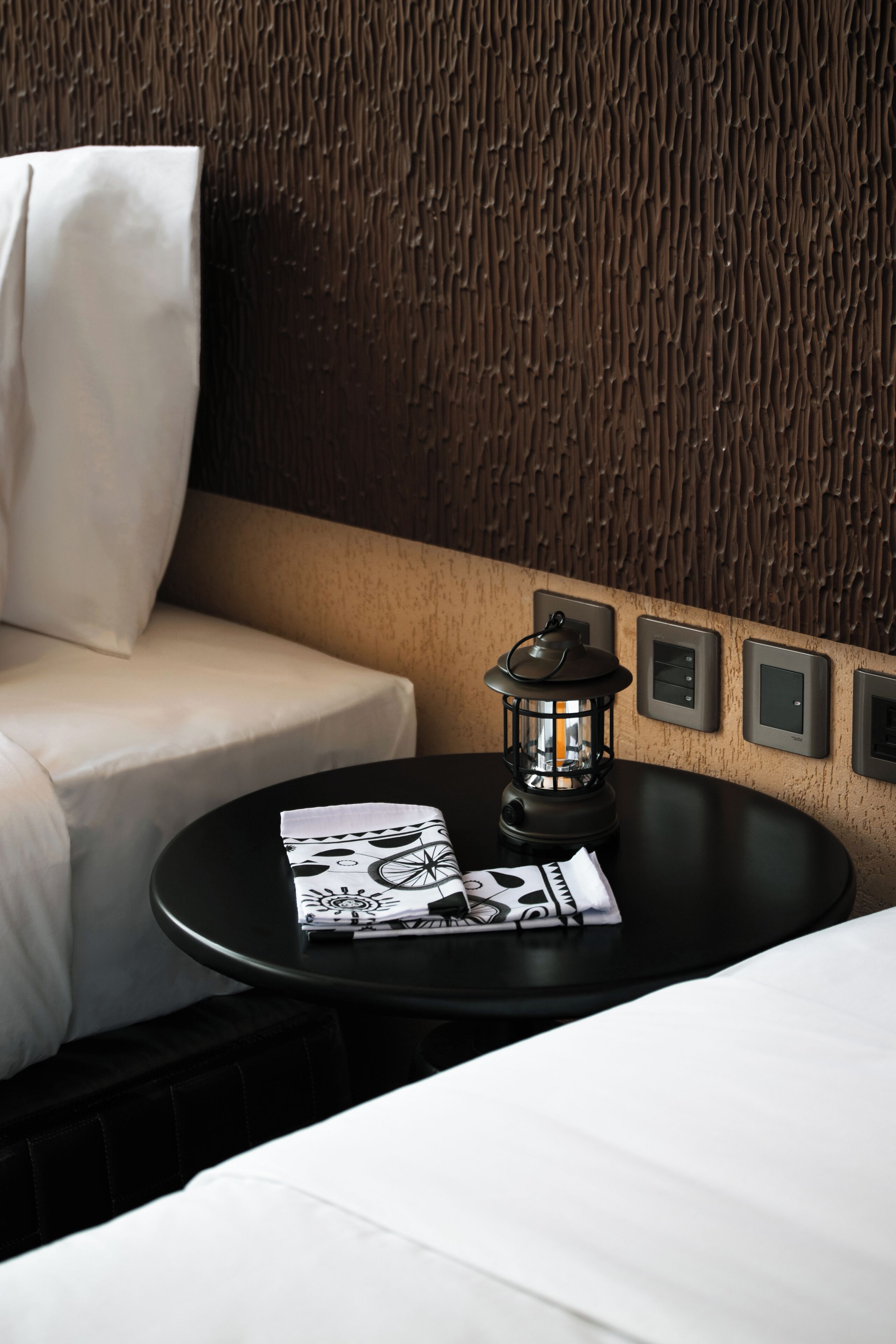 A modern hotel room with a textured wall, a black round table with a lantern, and a white sofa.