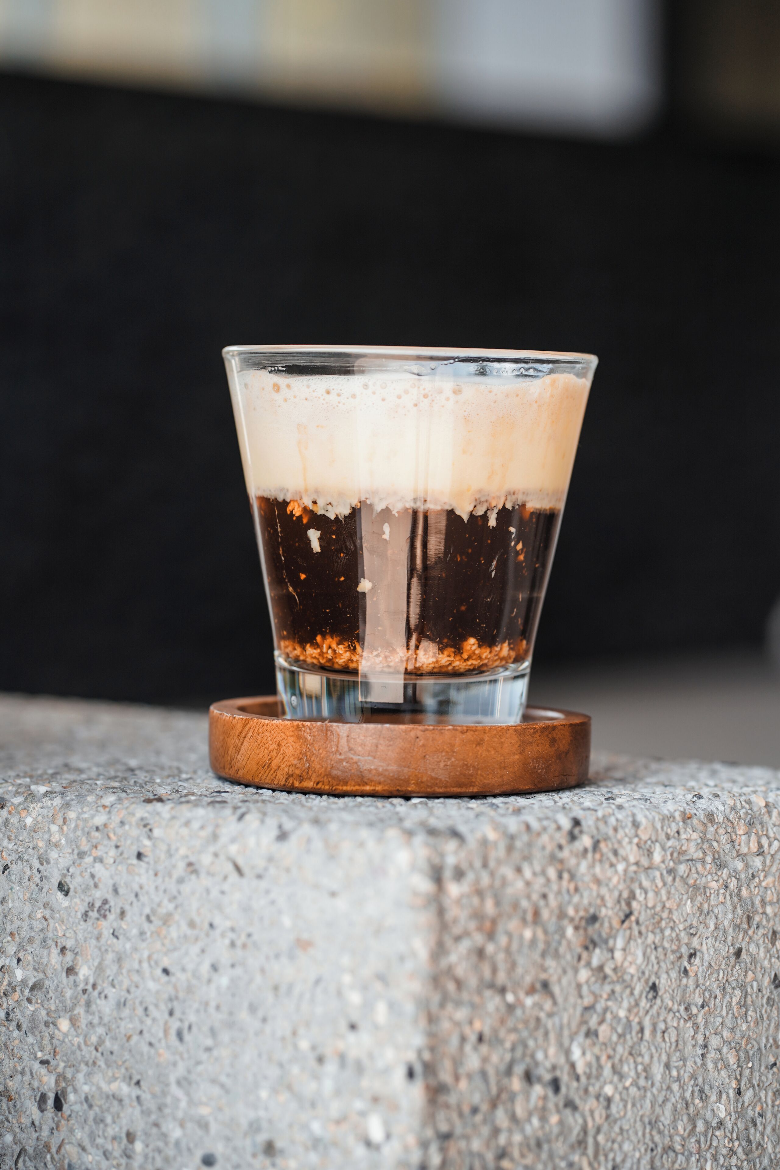 A glass of dark liquid with a foamy top, placed on a wooden coaster on a concrete surface.
