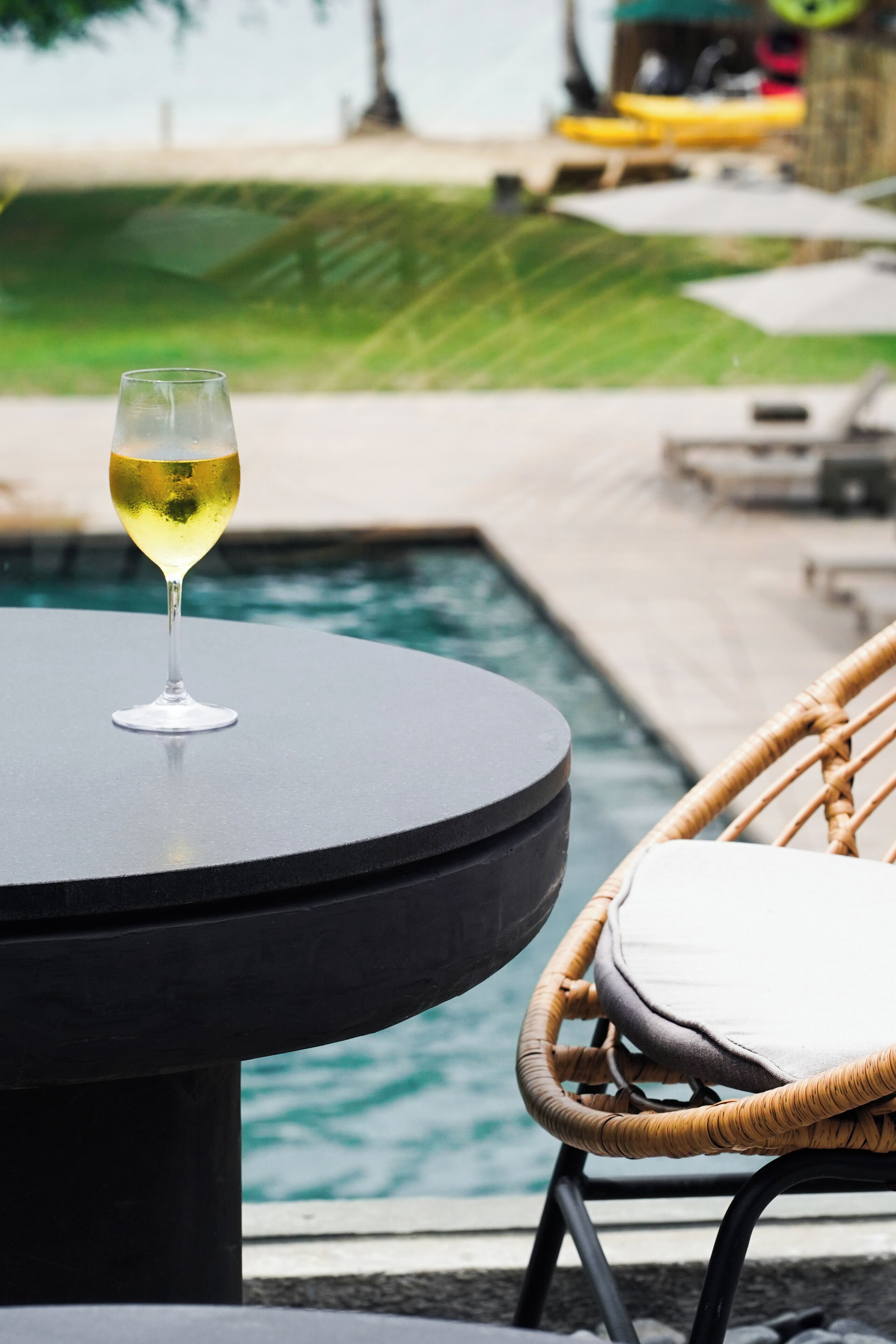 Un tavolo rotondo con un bicchiere di vino bianco, una sedia in vimini con un cuscino e una piscina sullo sfondo.