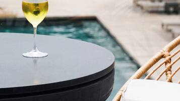 Un tavolo rotondo con un bicchiere di vino bianco, una sedia in vimini con un cuscino e una piscina sullo sfondo.
