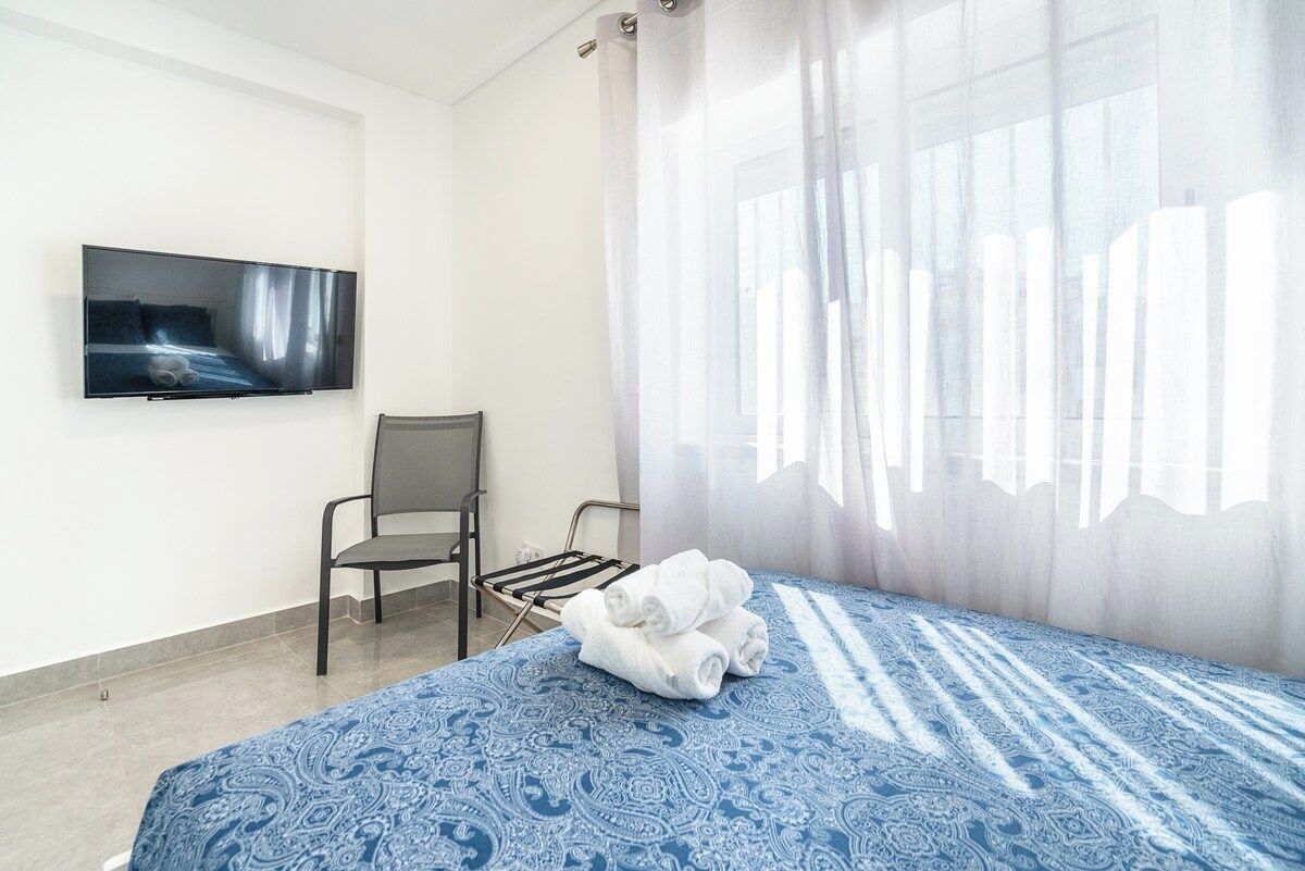 Ein Hotelzimmer mit Bett, einem Sessel, einem an der Wand befestigten Fernseher und einem Fenster mit durchscheinenden Vorhängen.