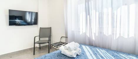 Una habitación de hotel con una cama, una silla, un televisor montado en la pared y un ventanal con cortinas translúcidas.