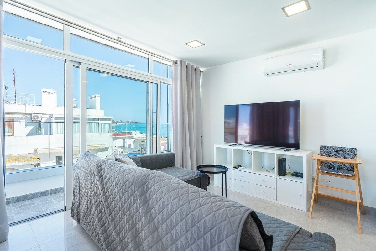 A modern living room with a large flat-screen TV, a sofa, a small table, and a balcony with a view of the sea.