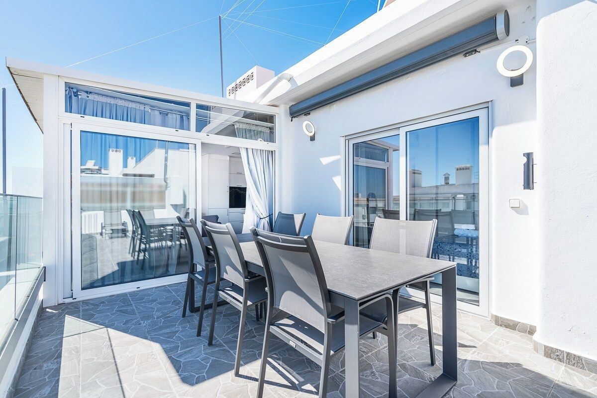 Un área de comedor moderna al aire libre con mesa y sillas, un ventanal de vidrio que da a un espacio interior, y vista panorámica de la ciudad.