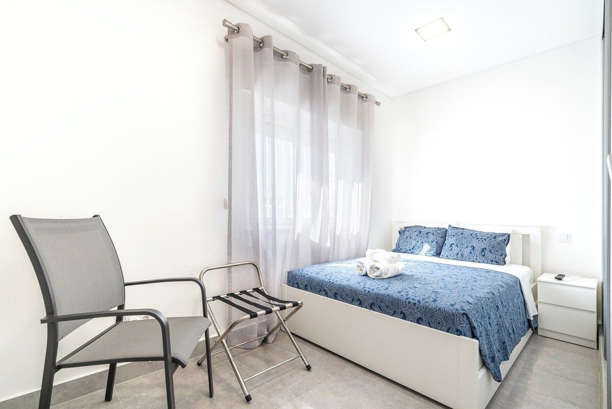 Un dormitorio con cama, una silla, una mesita de noche y una ventana con cortinas traslúcidas.