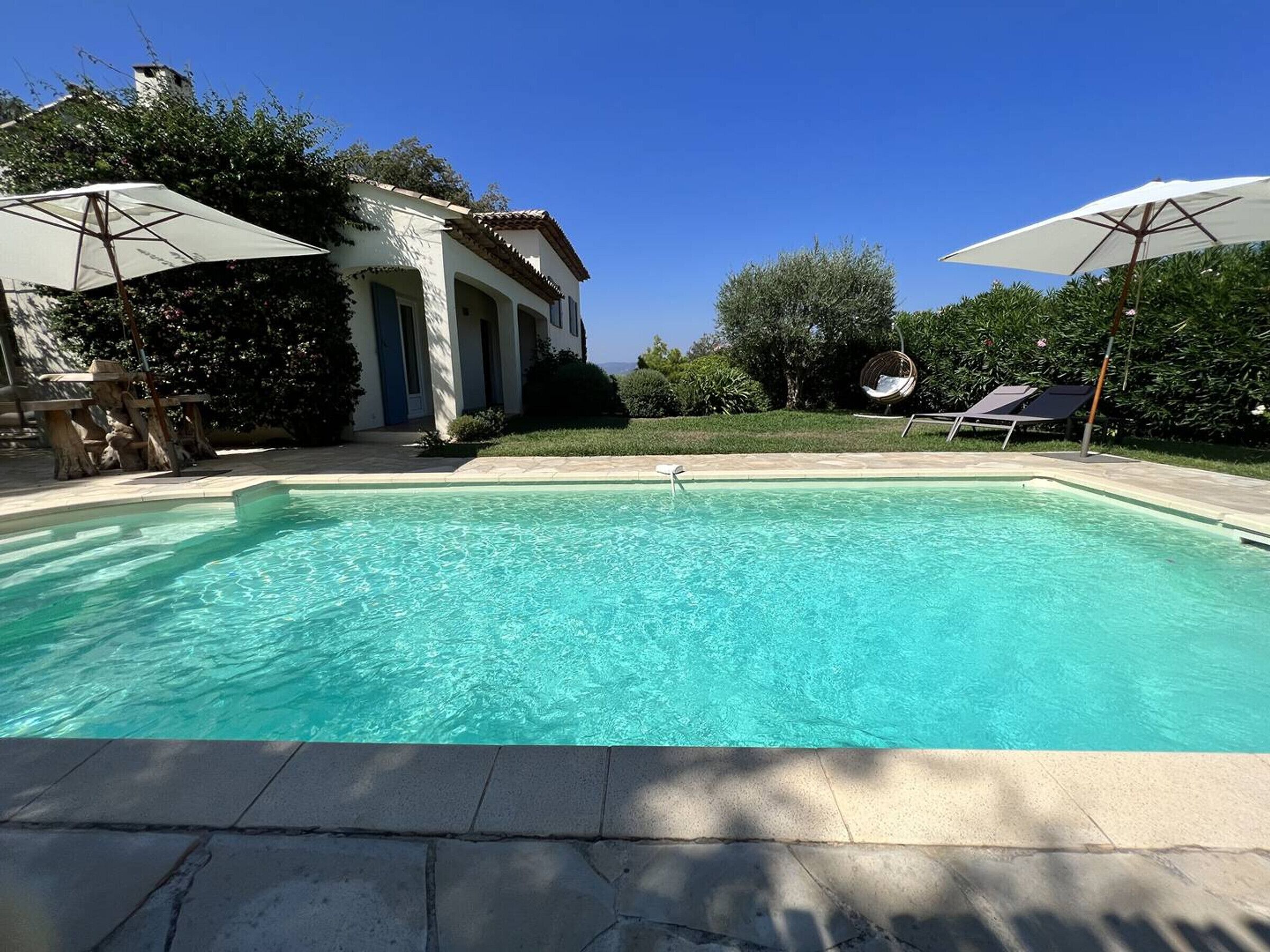 Une piscine à l’eau d’un bleu limpide, entourée d’une terrasse en pierre. Deux parasols offrent de l’ombre, et on aperçoit une chaise suspendue à l’arrière-plan.