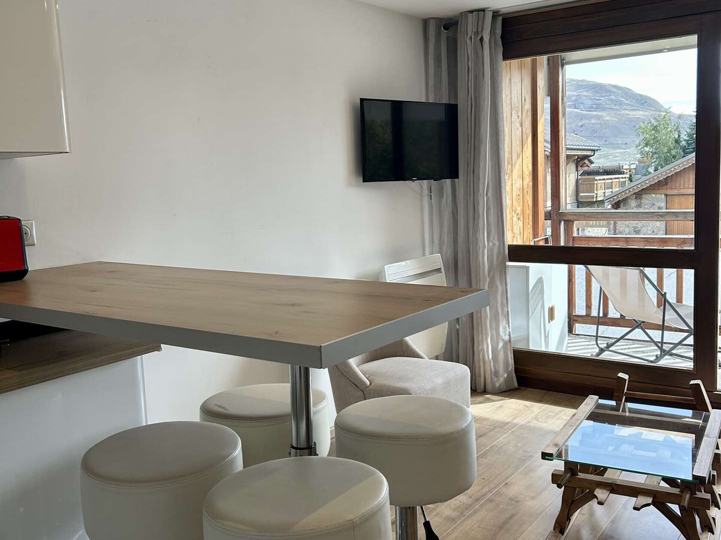 Ein modernes Zimmer mit einem Holzespeisetisch, weiß gepolsterten Sitzgelegenheiten, einem flachbildfernseher an der Wand und Blick auf eine gebirgige Landschaft durch große Fenster.