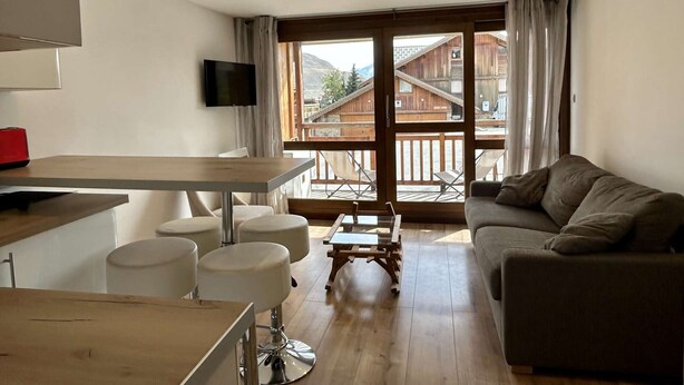 A modern living room with a sofa, a dining table with chairs, a TV mounted on the wall, and a balcony with wooden railings.