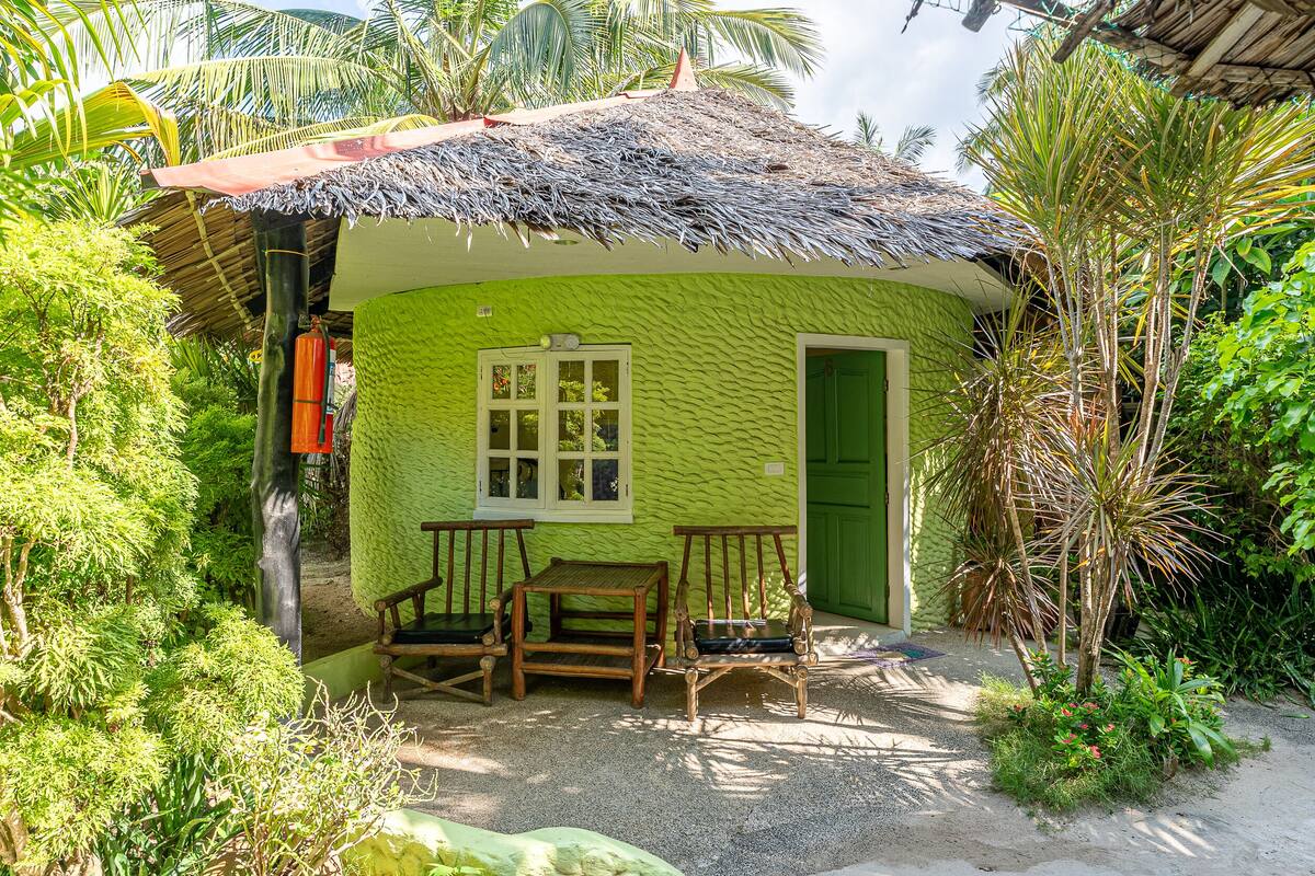 A small, green house with a thatched roof, a wooden table, two wooden chairs, and a green door.