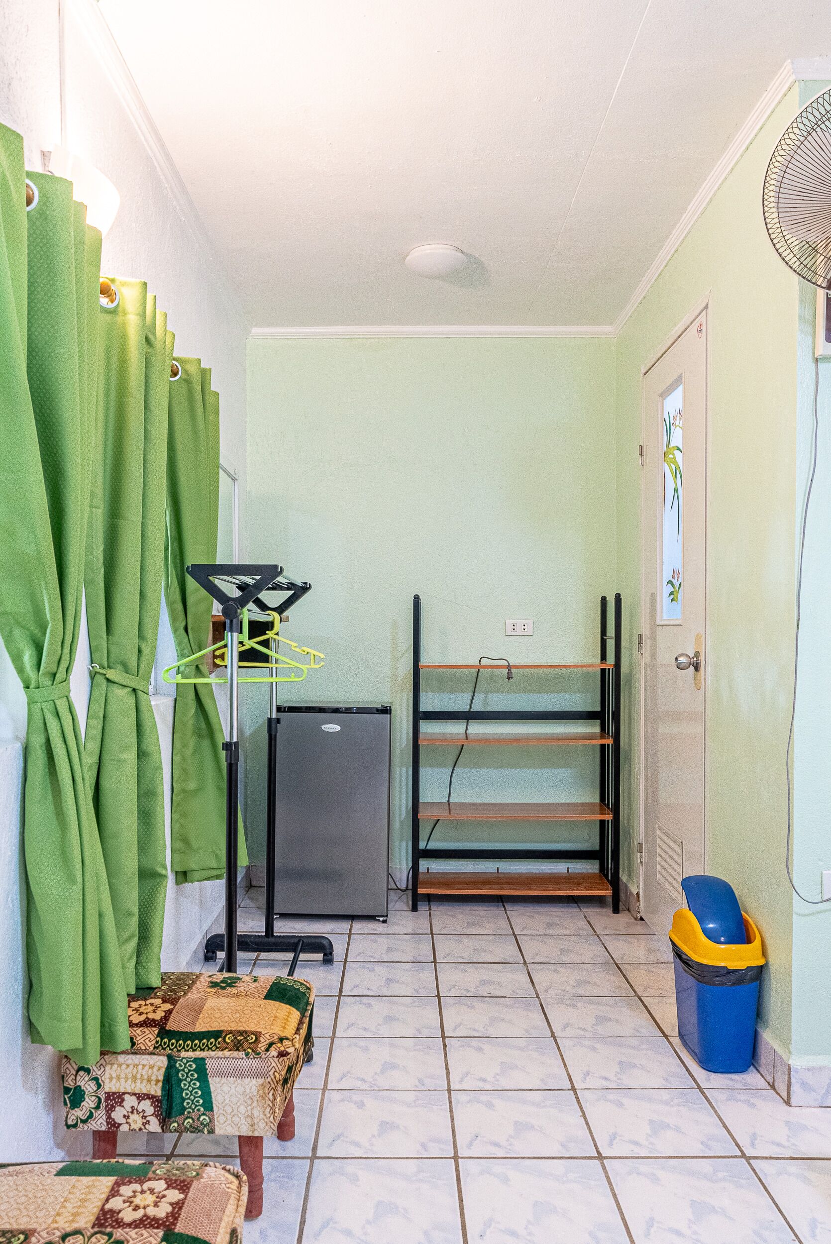 Una habitación pequeña con suelo de baldosas, un refrigerador, un ventilador, una puerta con un adhesivo decorativo y un cubo de basura.