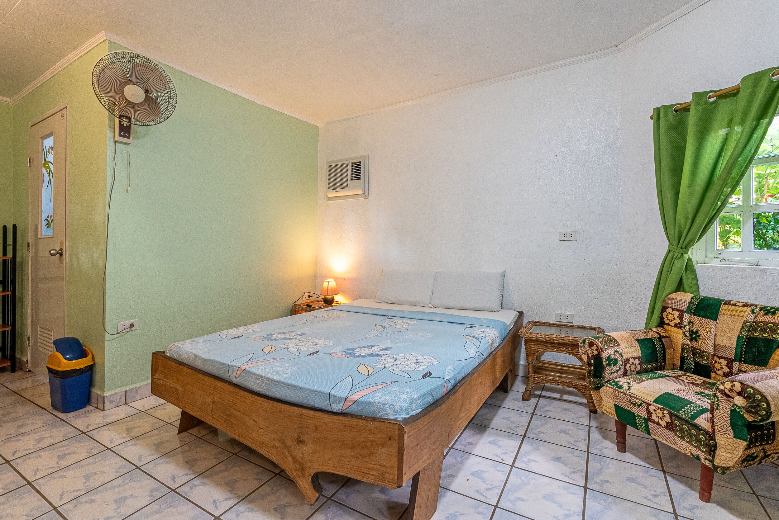 A room with a wooden bed, a small bedside table with a lamp, a wicker chair, and a fan on the wall.