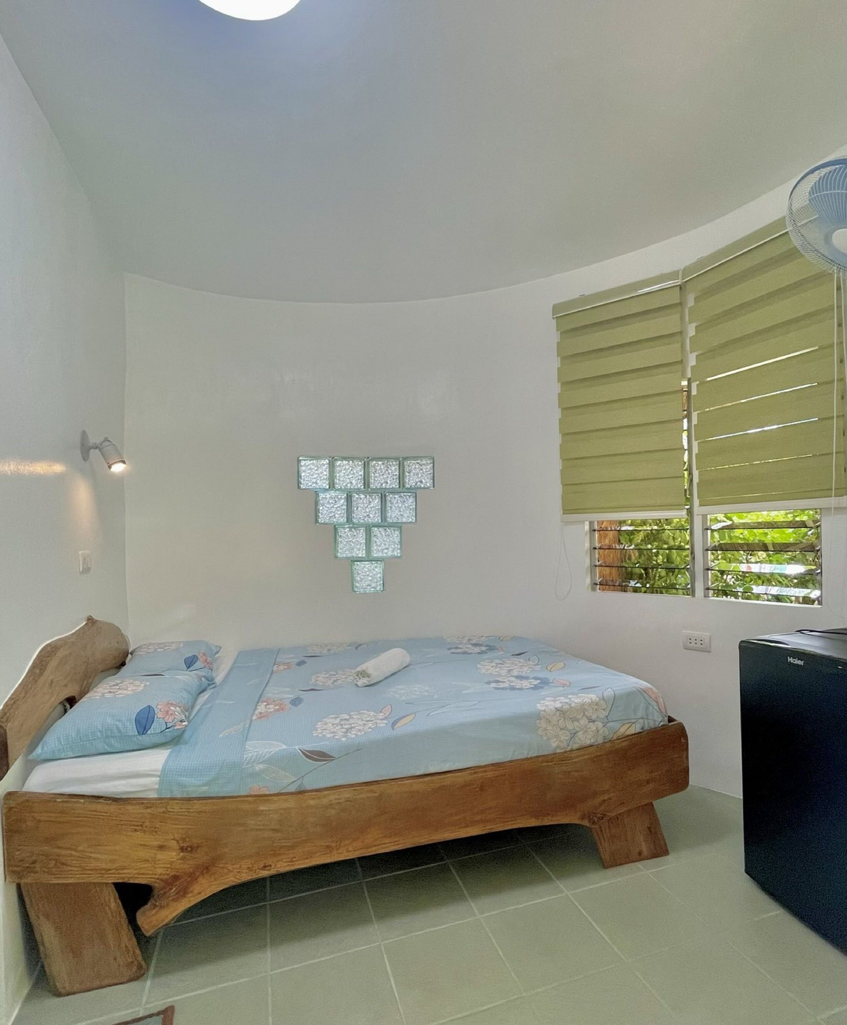 Quarto com cama de madeira, uma pequena escrivaninha, luminária fixada na parede e janela com persianas.