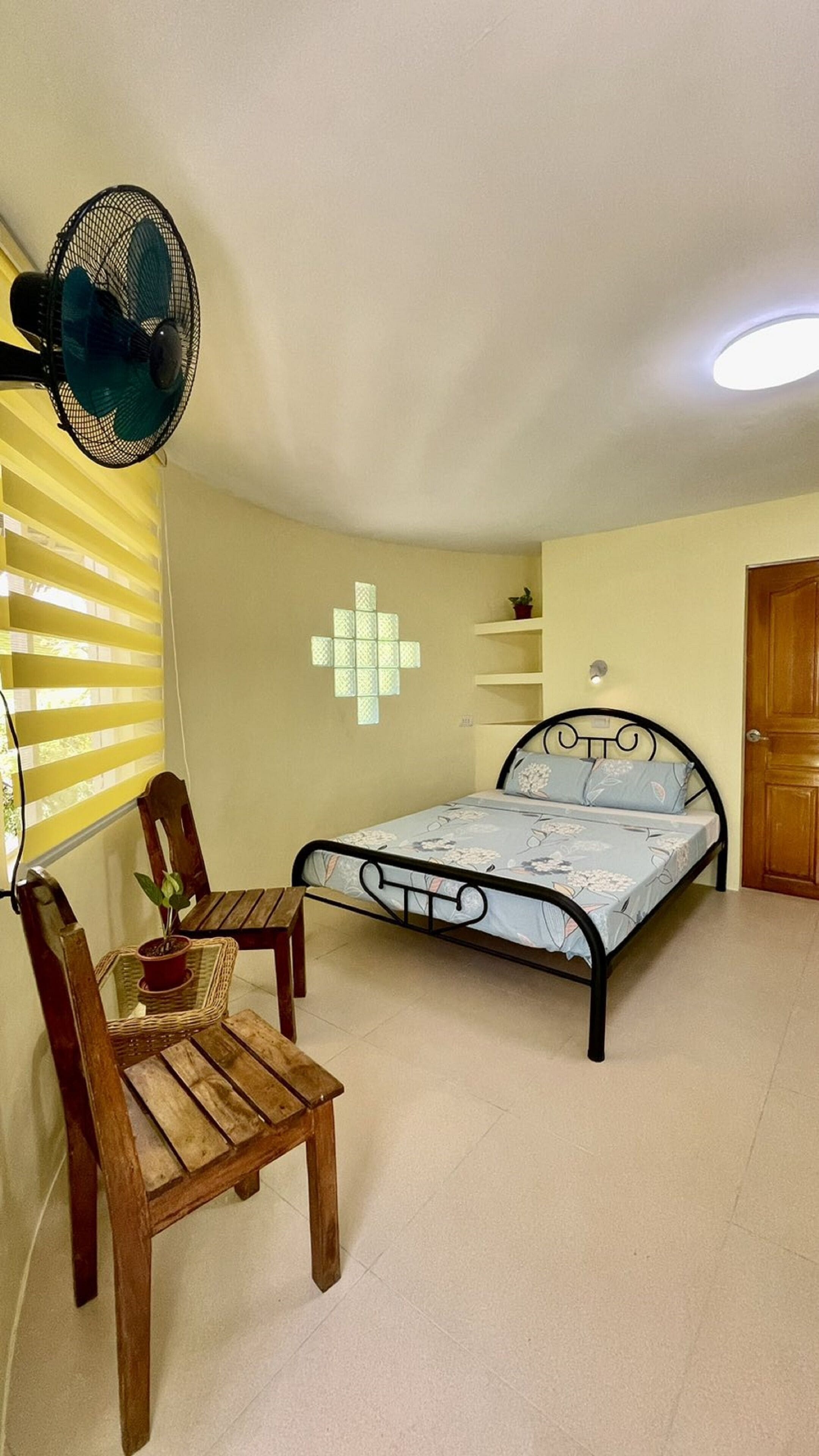 Un dormitorio con cama, una silla de madera, una mesita, un ventilador de pared, una ventana con persianas, un estante con una planta y una puerta.