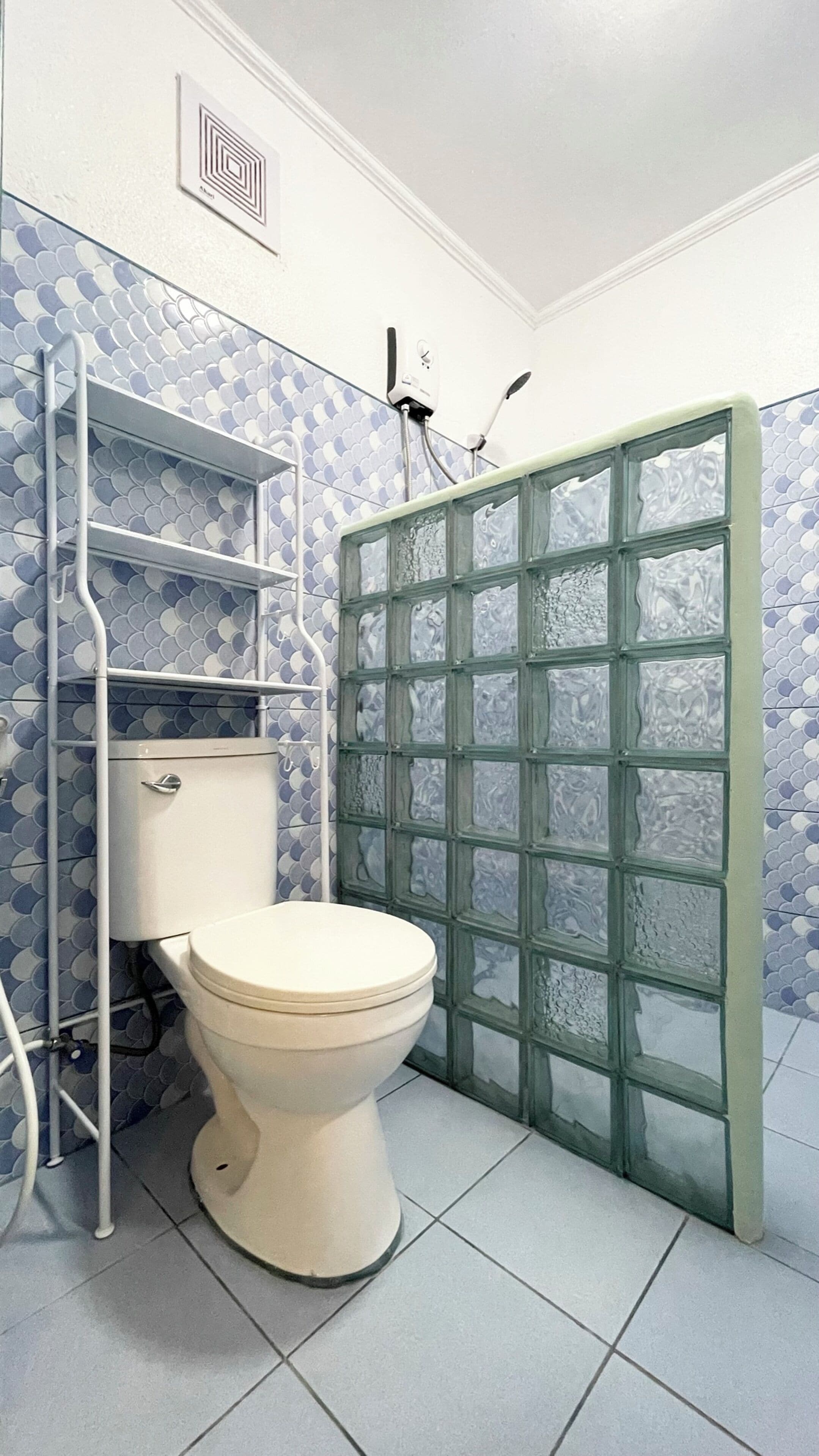 Un baño con inodoro, una estantería metálica, cabina de ducha con paneles de vidrio esmerilado y un salpicadero de azulejos estampados.