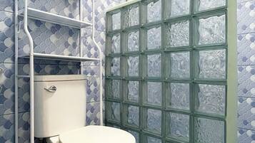 Un baño con inodoro, una estantería metálica, cabina de ducha con paneles de vidrio esmerilado y un salpicadero de azulejos estampados.