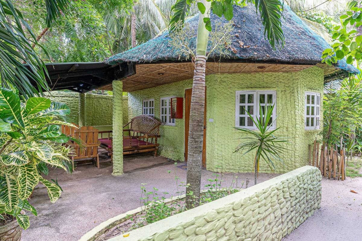 A small, single-story house with a thatched roof, surrounded by lush greenery.