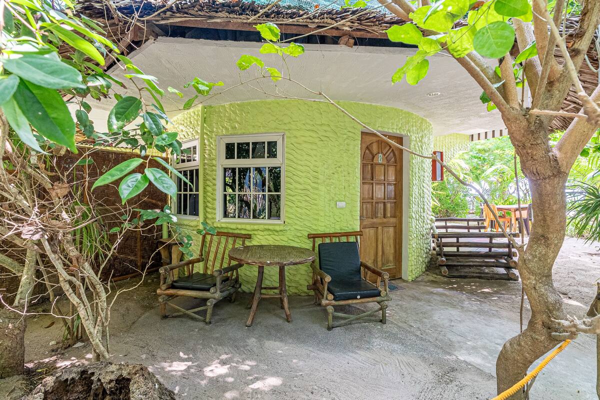 A green building with a window and door, surrounded by trees and a table with chairs.