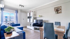A modern living room with a sofa, a dining table with chairs, a TV, and a small plant on a side table. - Beachfront Apartment 4F - Armação de Pêra, Algarve (Armação)