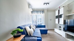 A modern living room with a blue sofa, a small table with a plant, and a TV on a stand. - Beachfront Apartment 4F - Armação de Pêra, Algarve (Armação)